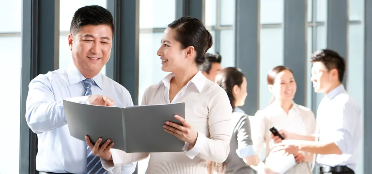 Two business professionals reviewing a document, with colleagues chatting in the background