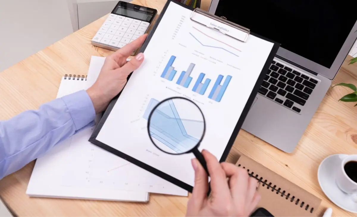 Hands with magnifying glass over sales charts on clipboard.